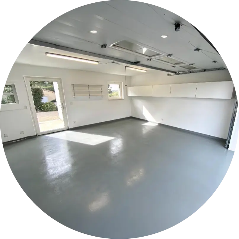 Vue intérieure d'un garage moderne à Antibes avec un sol en résine époxy gris brillant, des rangements blancs épurés et une porte ouverte laissant entrer la lumière naturelle.